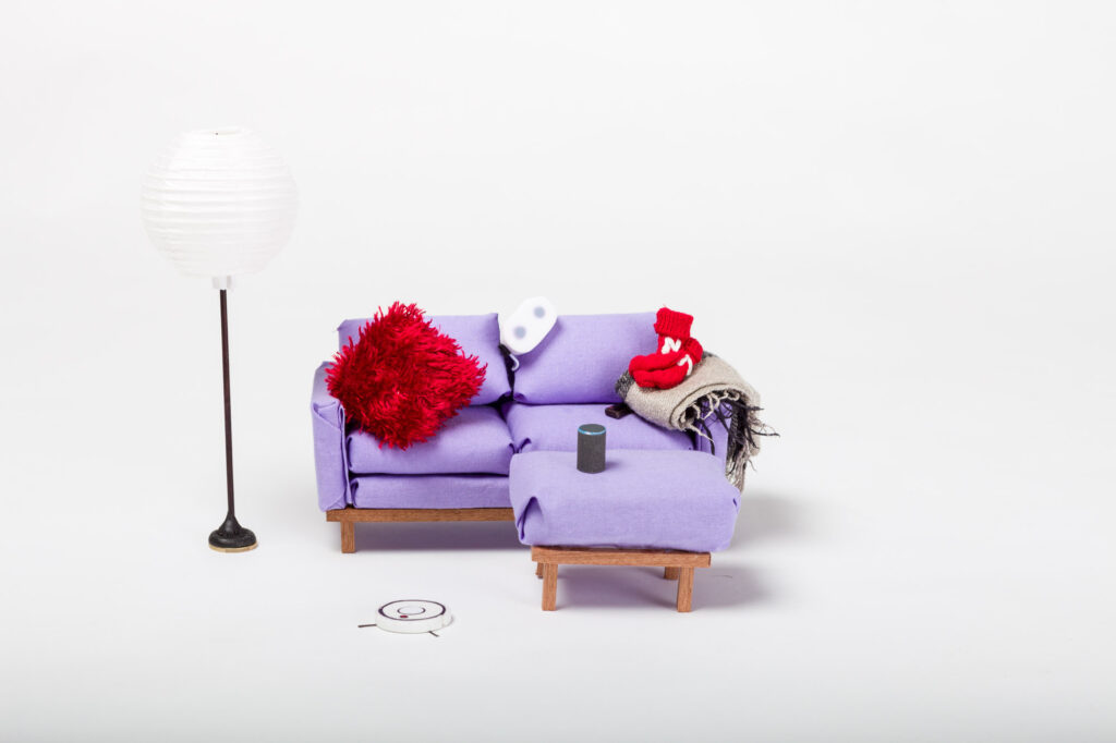 Un juego de sofá en miniatura. En el sofa hay unas medias rojas, un casco de realidad virtual y una Alexa. Frente al sofá hay un Roomba